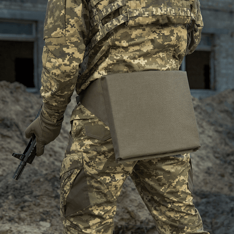 M-Tac sitteunderlag med belte i bruk av en soldat i kamuflasjeuniform, sett bakfra på en jordet grunn.