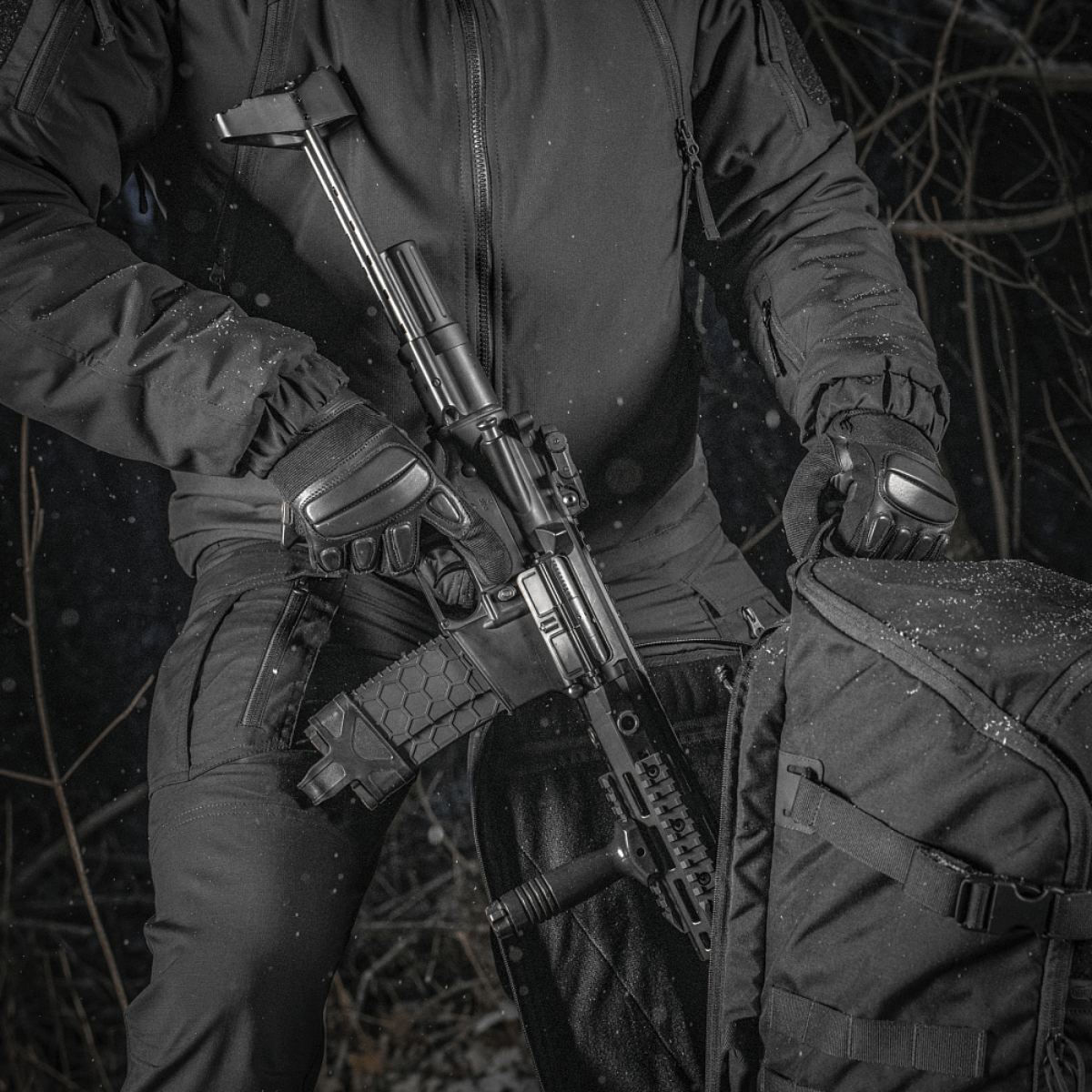 En person holder et våben ved siden af en M-Tac Rygsæk Gen.II Elite Large i en skovområde. Rygsækkens holdbare design er tydeligt.