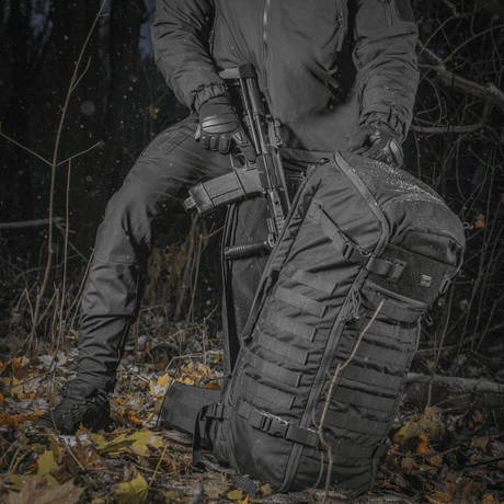 Person med M-Tac Ryggsekk Gen.II Elite Large i skog, med våpen for transport, slitesterkt Cordura-materiale og ergonomisk design.