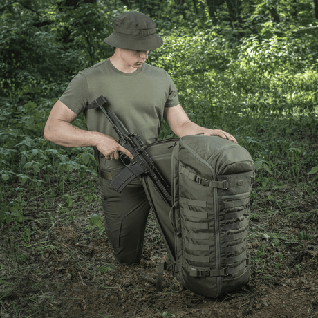 M-Tac Ryggsekk Gen.II Elite Large i skogen med våpen, viser holdbar konstruksjon og funksjonalitet for utendørs eventyr.