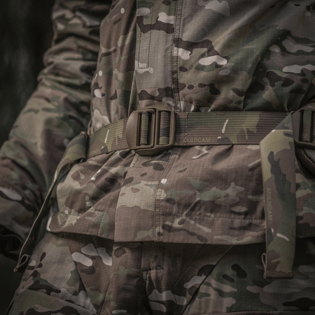 Nærbilde av militær uniform med kamuflasjemønster, fokus på belteområdet i skogmiljø.