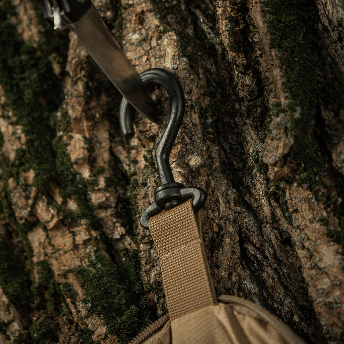 M-Tac Toilettaske Elite Gen.II hænges på en træstamme med en metalkrog, perfekt til organisering af rejseartikler og hygiejneprodukter.