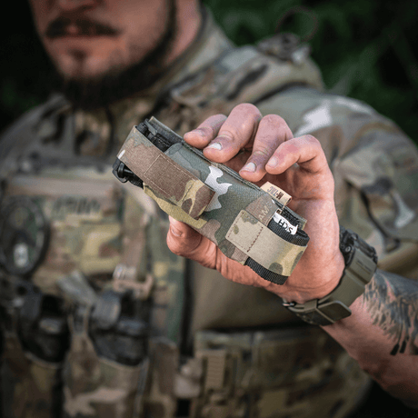 M-Tac Tourniquet Pouch Elastisk med MOLLE Gen.II i kamuflasje, holdt av person i militæruniform, lomme for rask tilgang til tourniquet.