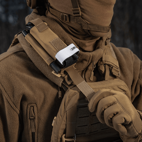 Person med brun bekledning bærer M-Tac Elastisk Tourniquet Pouch Molle Gen.III festet på vest, utstyrt med taktisk utstyr.
