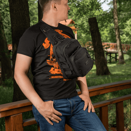 Mann bærer M‑Tac Cross Bag Elite Hex slingveske på en bro i skogen, perfekt for daglig bruk. Vesken rommer opptil 5 liter.