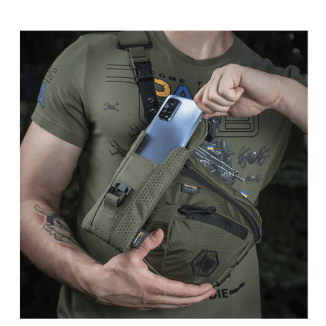 Mann med M-Tac Smartphone Lomme Elite Large Hex i mørkegrønn farge holder en smarttelefon.