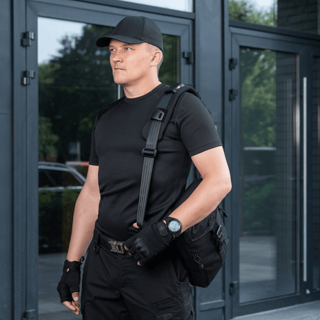 Mann bærer veske med M‑Tac skulderrempute 50 mm Elite for økt komfort og støtte, kledd i svart foran moderne bygning.
