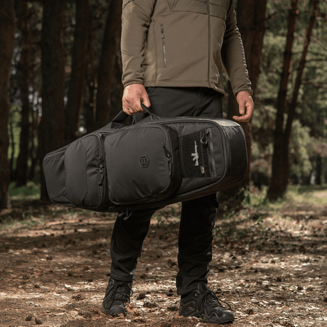 Mand bærer M‑Tac Gun Backpack Case 105 cm Elite Hex i skov, ideal til jagt og skydning, designet til AR-15 og AK karbiner.