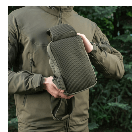Person viser M-Tac Sling pistolveske Elite Hex utendørs i skogmiljø, iført jakke med glidelås.
