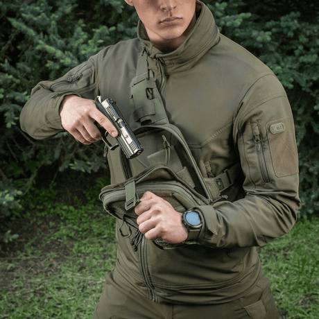 Person med M-Tac Sling pistolveske Elite Hex bærer en pistol foran et skogkledd bakteppe.