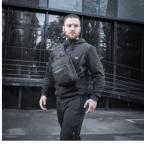 Mann med M-Tac Cross Taske Slim Elite Hex iført sorte klær foran moderne bygning.