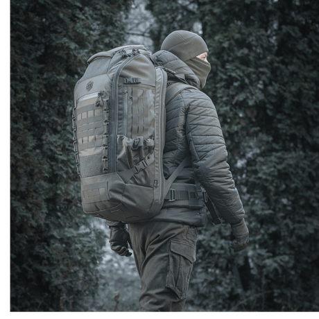M-Tac Ryggsekk Large Elite Hex Gen. IV i et skogmiljø, designet for bæring av våpen og beskyttelse med slitesterkt Cordura-materiale.