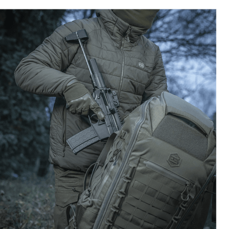 Person bærer våpen ved siden av M-Tac Ryggsekk Large Elite Hex Gen. IV i naturen, viser sekkens holdbarhet og design.