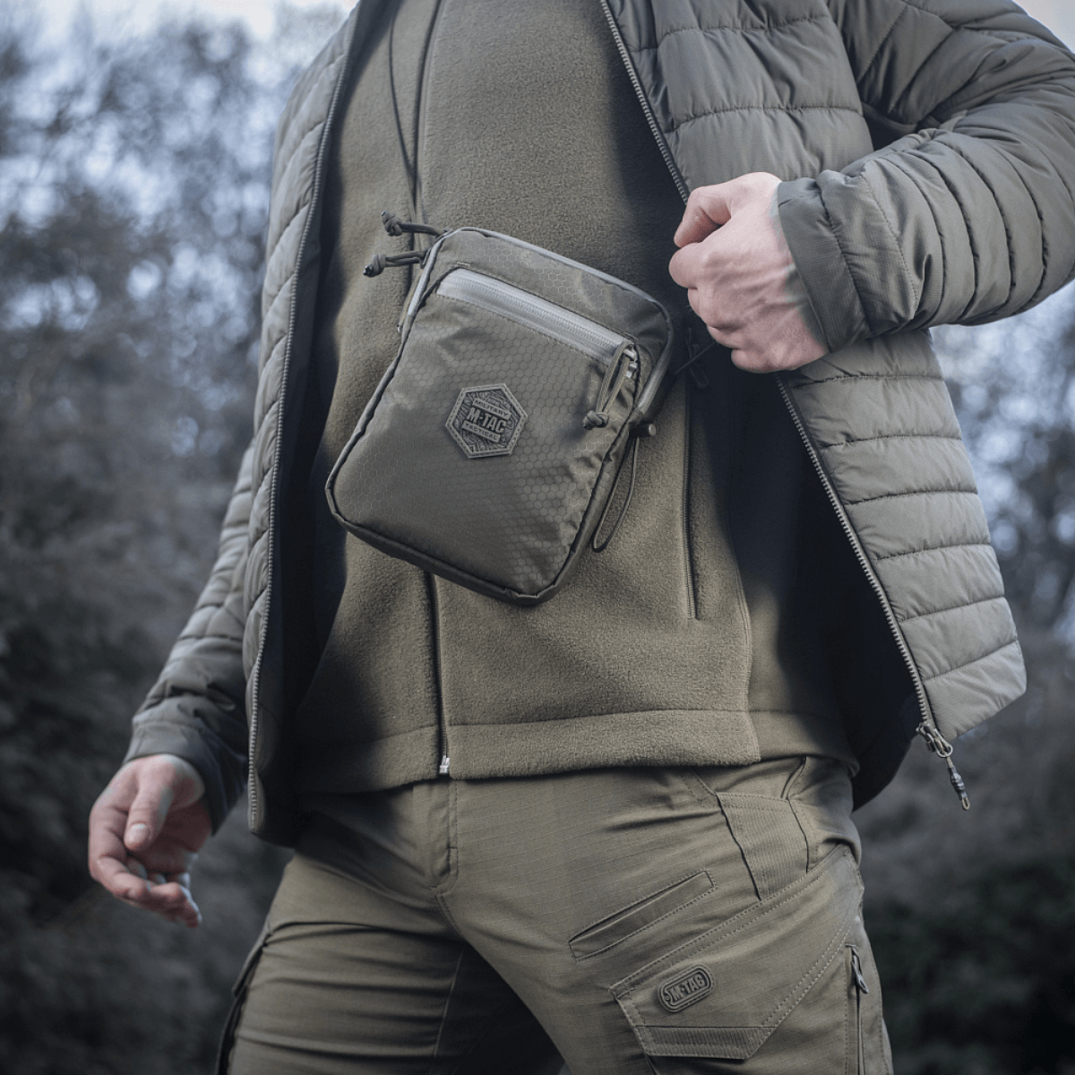 Person wearing the M‑Tac Pocket Bag Elite HEX in a rugged outdoor setting, showcasing its portability and durability.