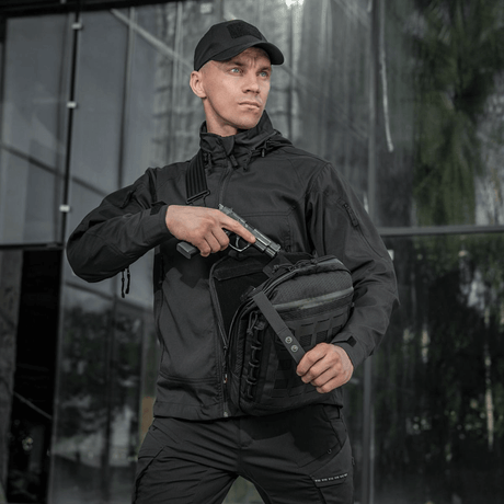 Person holder M-Tac Tablet Bag Laser Cut Hex og avdekker skjult våpen foran glassfasade, iført svart jakke og lue.