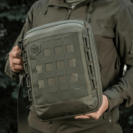 M-Tac Tablet Bag Laser Cut Hex med Laser Cut MOLLE-panel, laget av Cordura-materialer, holdt av person i friluftsmiljø.