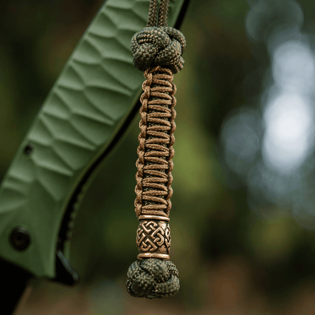 M-Tac Knife Lanyard Celtic i flettet paracord med dekorativt keltisk mønster, festet til grepsdelen på en kniv.