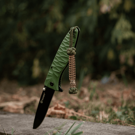 M-Tac Knife Lanyard Celtic - grønn foldekniv med flettet paracord-lanyard i utendørs miljø.