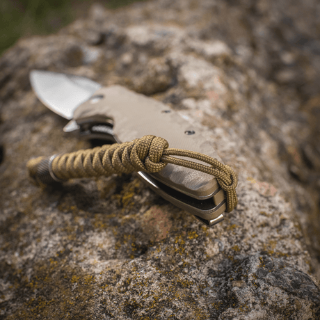 M-Tac Viper Lanyard Stainless Steel festet til kniv, vist på steinoverflate. Fleksibel forlengerdel i flettet paracord.