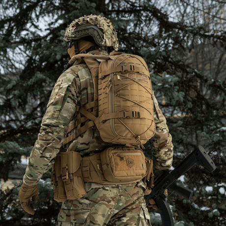 M-Tac ryggsekk Sturm Elite Gen.II i feltet, mann i kamuflasje og taktisk ryggsekk med MOLLE-system og forsterkede stropper, skogmiljø.