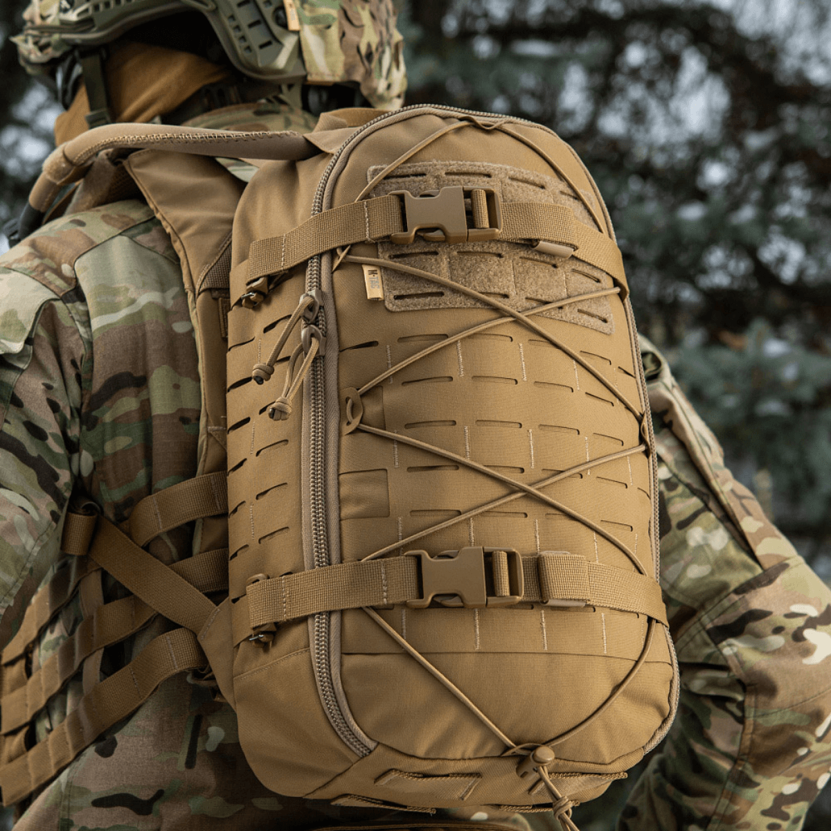 Soldat bærer M-Tac Rygsæk Sturm Elite Gen.II i ørkenfarve, med elastisk snøring, MOLLE system og Cordura konstruktion, i feltmiljø.