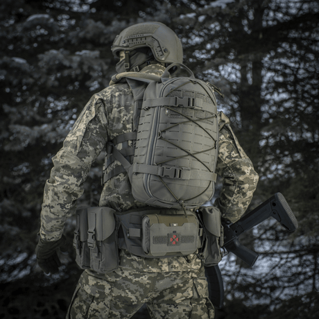 Taktisk person med M-Tac ryggsekk Sturm Elite Gen.II i skogmiljø iført kamuflasjeuniform, sett bakfra.