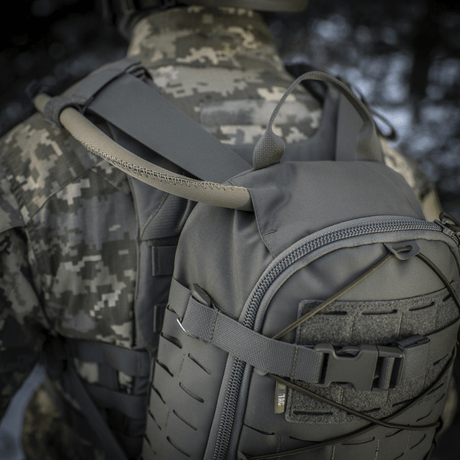 M-Tac ryggsekk Sturm Elite Gen.II i grå Cordura på en soldat i kamuflasje, viser integrert drikkesystem med slange i naturen.