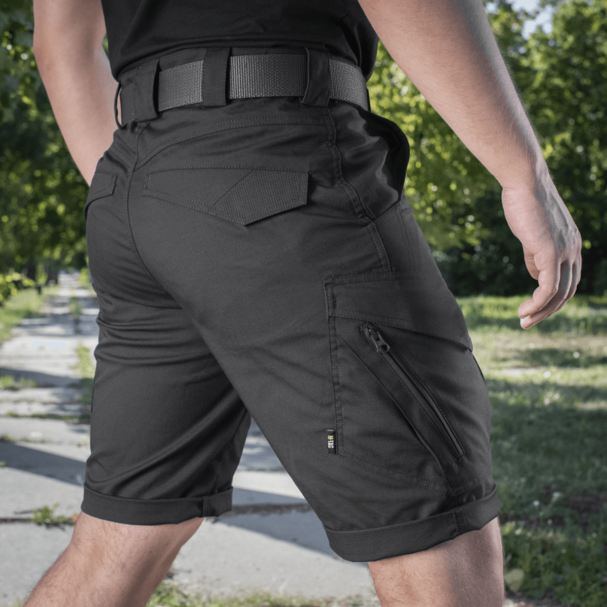 M-Tac Shorts Aggressor Gen.II Flex til alsidig brug udendørs, sort design, mand i shorts på gåtur i parkområde, let og funktionel pasform.