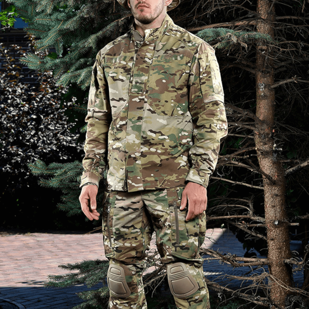 Person iført militæruniform i camouflage, står blandt træer, klar til outdoor aktiviteter.