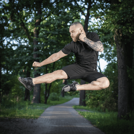 Mand i M-Tac Shorts Casual Fit Cotton udfører et spring i parken, iført afslappet sort tøj, fremstillet af åndbart 100% bomuld.