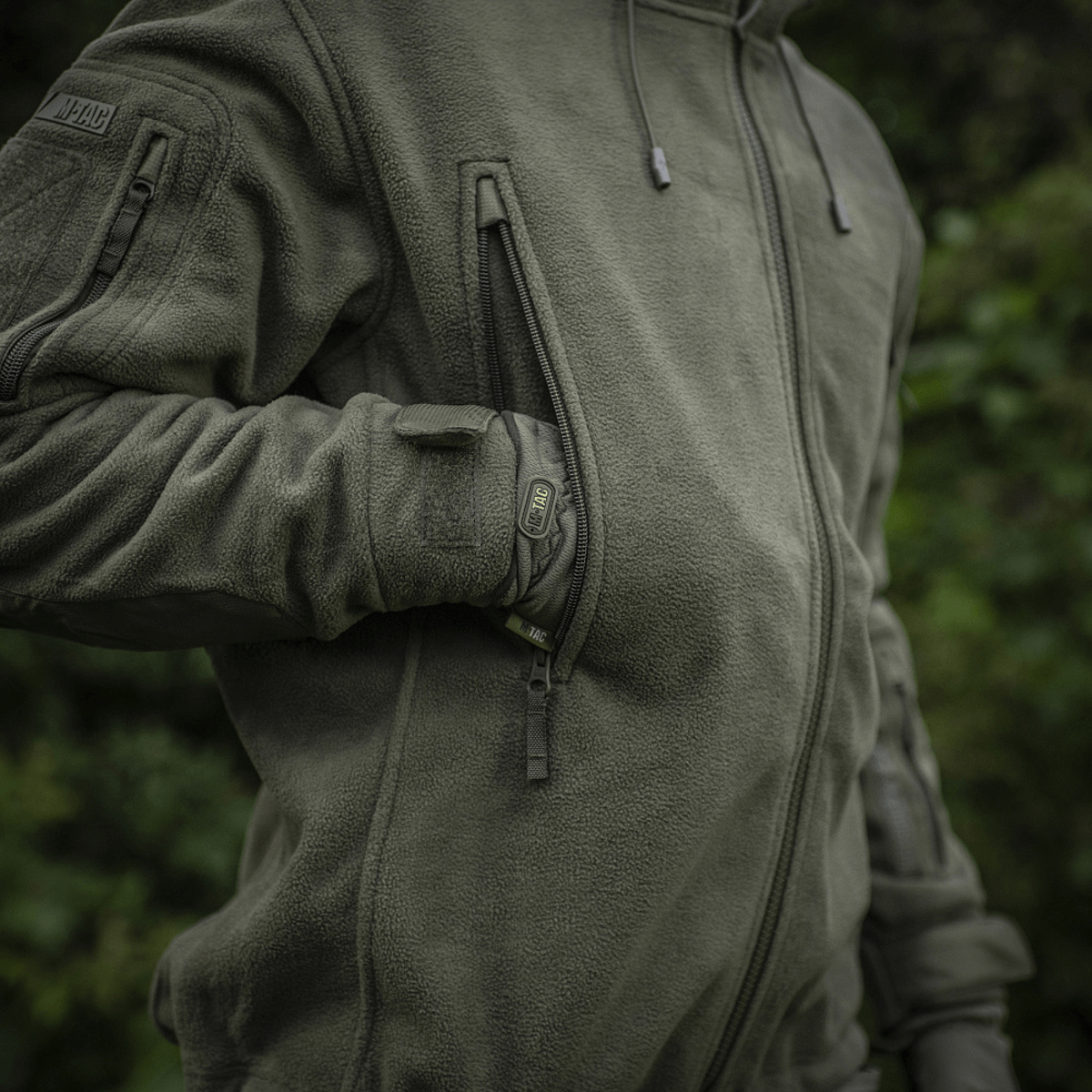 En person iført M-Tac Division Gen.II Windblock Fleecejakke, designet til maksimal varme og vindbeskyttelse, set i grøn skovbaggrund.