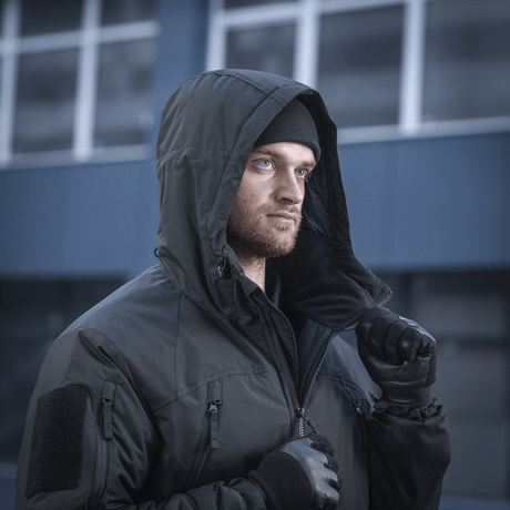 Mand iført M-Tac Alpha Gen.III Pro Vinterjakke med hætte oppe, komfortabel ved temperaturer ned til -20° C, på baggrund af moderne bygning.
