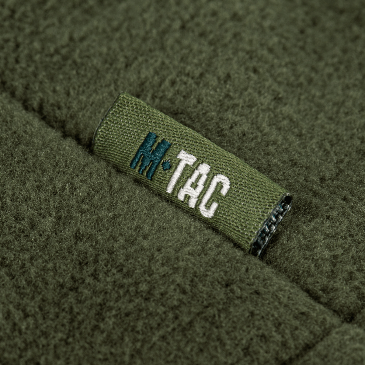 Detalje af M-Tac Combat Fleece Polartec Jakke med mærkelogo i olivengrøn farve, der viser høj kvalitet og avanceret design.