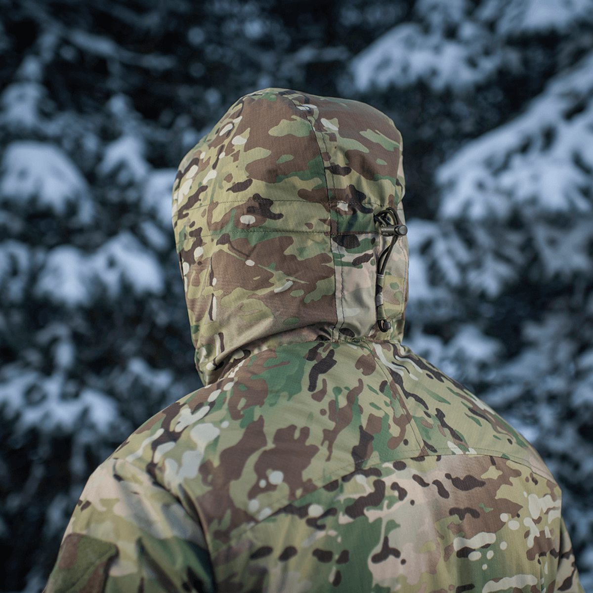 M-Tac Alpha Gen.IV Pro Primaloft Vinterjakke i Multicam mønster med snebeklædte træer i baggrunden, optimal til -20°C.
