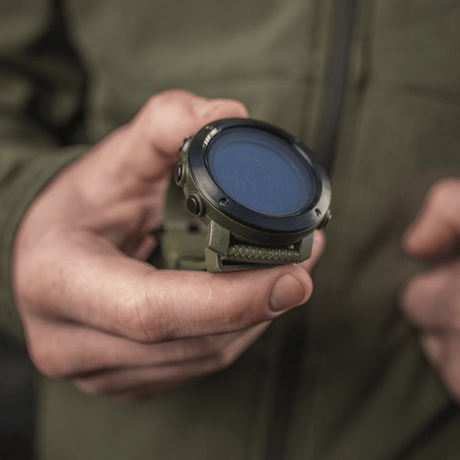M‑Tac multifunksjonelt taktisk ur holdt i hånden, ideelt for friluftsliv og militærbruk, viser robust design og avanserte funksjoner