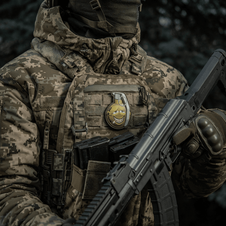 Soldat med kamuflasjejakke bærer M-Tac Patch M67 Granat 3D PVC på vesten, humoristisk emoji-design, taktisk militærutstyr.