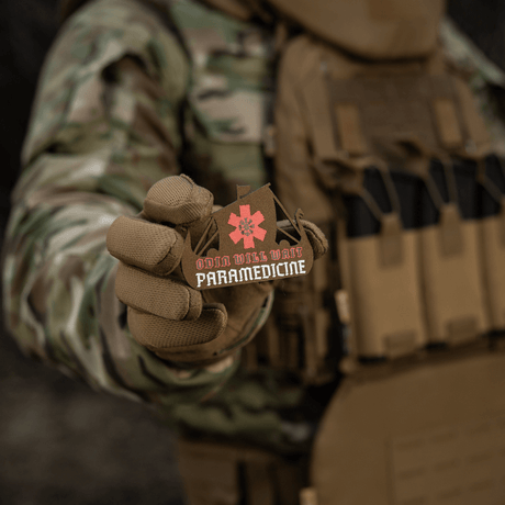 M-Tac Patch Viking Evac i soldats hånd, viser Odin will wait paramedicine-motiv. Kamuflasjejakke og taktisk vest i bakgrunnen.