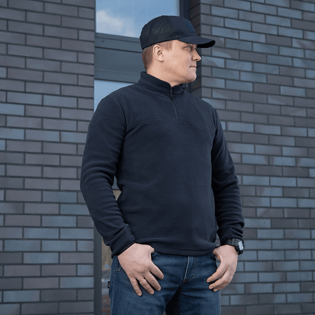 M-Tac Delta Polartec Fleecejakke i sort, fremstillet af Polartec®-fleece, bæres af en mand foran en murstensvæg.