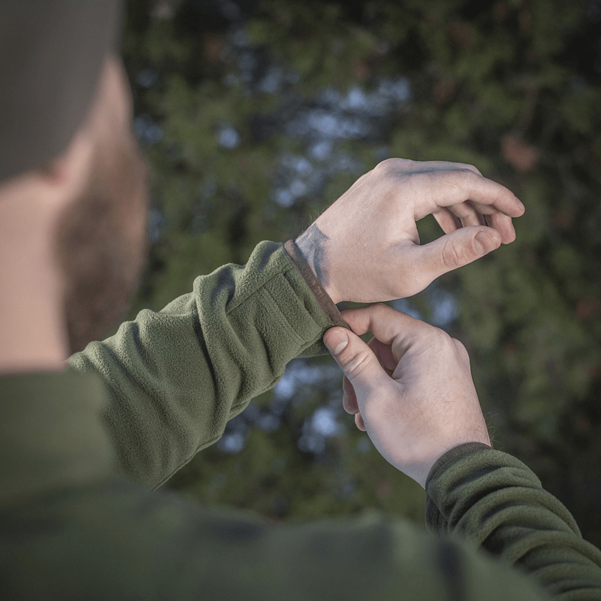 Mand justerer manchetten på en M-Tac Delta Polartec Fleecejakke i en skov, som viser jakkens funktionelle design og materiale.