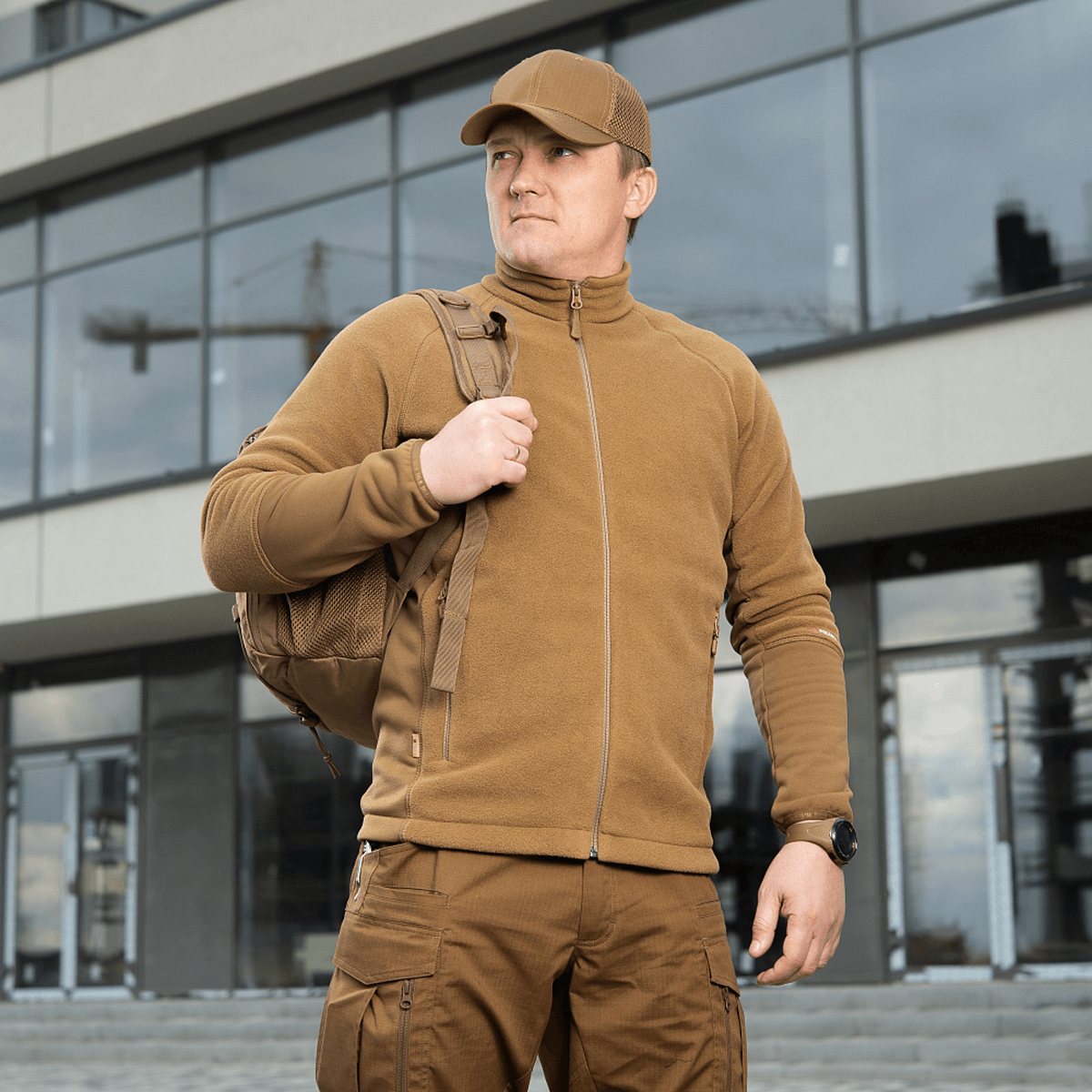 Mand iført M-Tac Polartec Sport Fleecejakke med rygsæk foran moderne bygning, viser funktionalitet og stil.