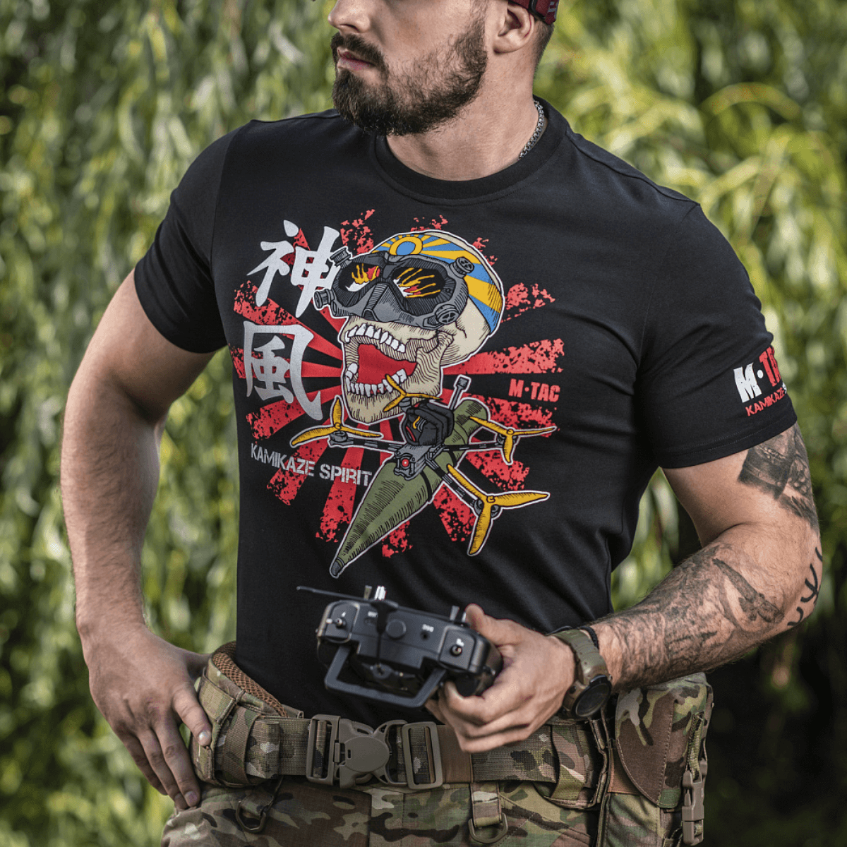 Mand iført M-Tac T-Shirt Kamikaze Spirit med stiliseret kranie-design og FPV-goggles, stående udendørs med en controller i hånden.