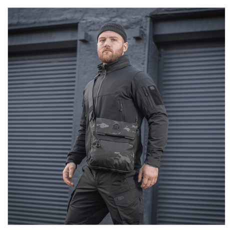 Mann iført M-Tac Konvert Taske Elite i svart camouflage-stil foran industrielle garasjeporter.
