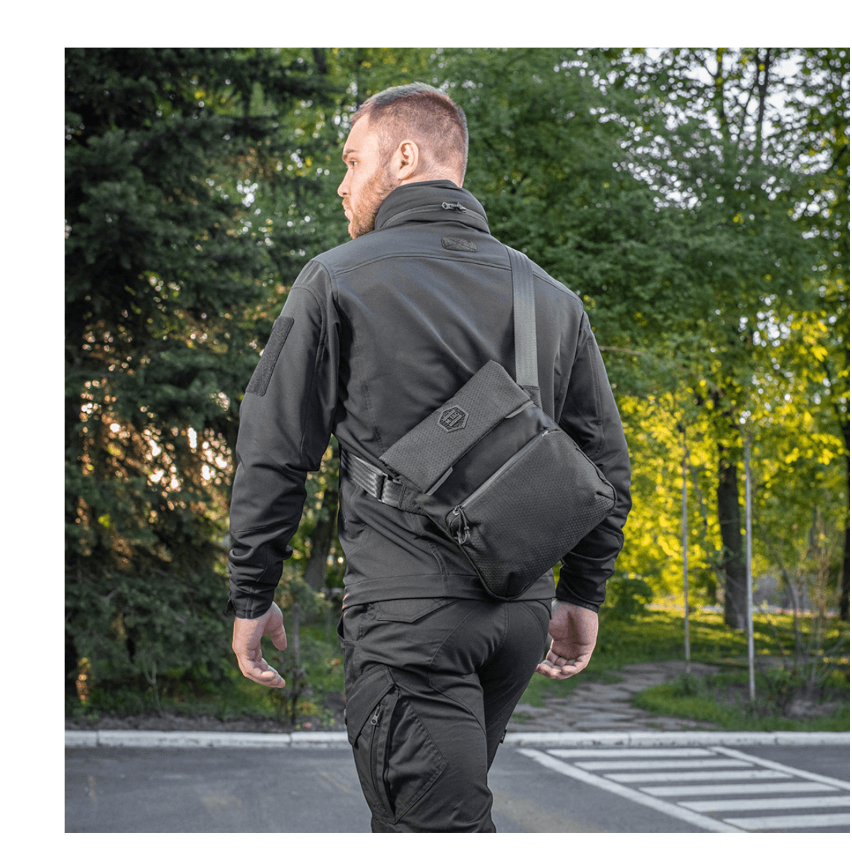 En mand bærer en M-Tac Konvert Taske Elite på ryggen i et udendørs miljø med træer i baggrunden.