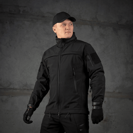 Mand iført M-Tac Soft Shell Police Jakke står klar til aktion, beskyttet mod vind og vejr med justerbar hætte.