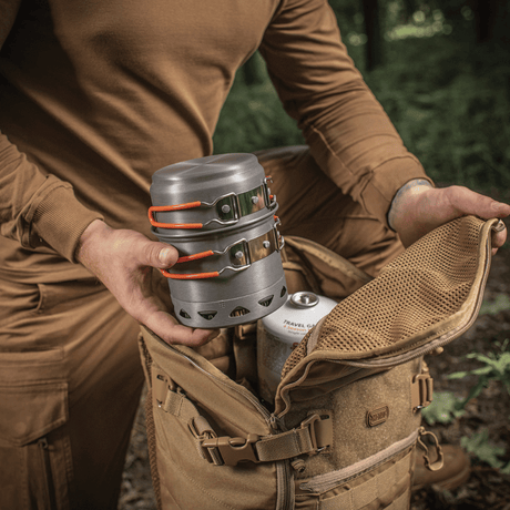 Person pakker M-Tac enkelt kokesett til brenner i ryggsekk i skogen, ideelt for friluftsliv og camping, laget av anodisert aluminium.