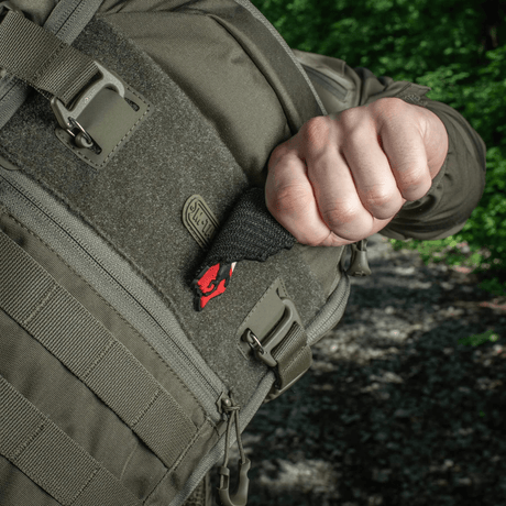 Person trekker en M-Tac Patch Husaria 3D PVC morale-patch ut av en taktisk ryggsekk, viser vinget husar-design mot grønn skogbakgrunn.