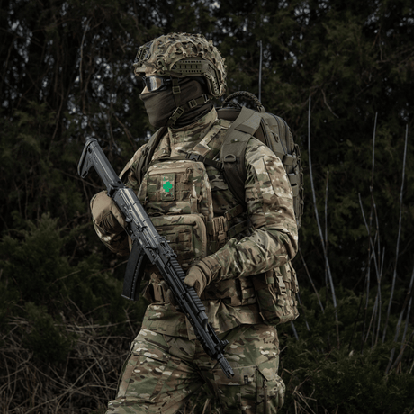 Soldat i militæruniform med M-Tac Patch Lucky Card (broderi) festet på vesten, symbol på hell og beskyttelse, kamuflasje i skogsområde.
