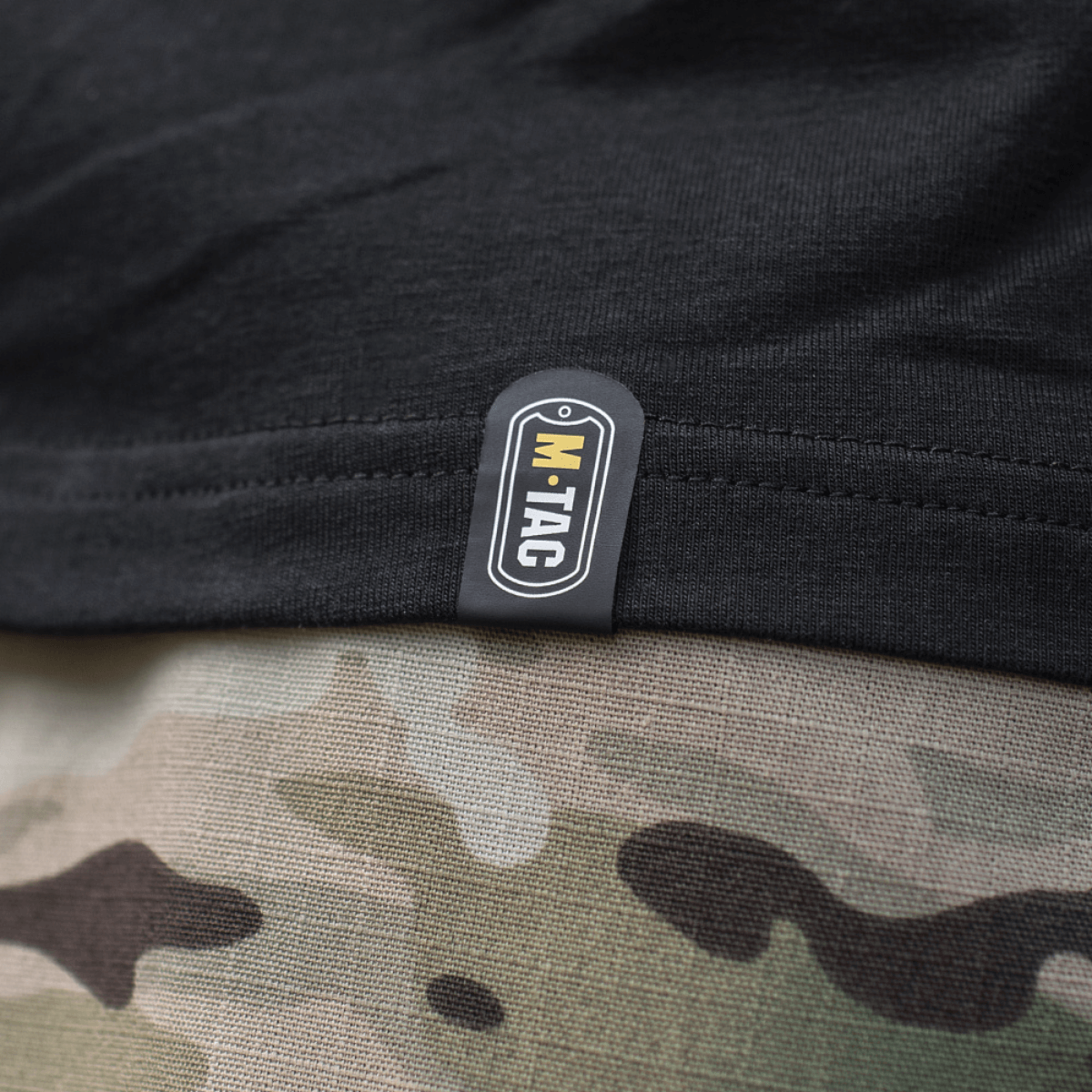 Nærbillede af M-Tac T-Shirt Surf Club tag, sort stof med kamuflagerprint i baggrunden