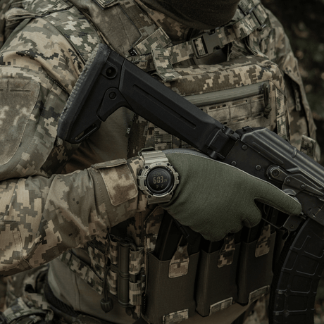 Person i kamuflasjeklær bærer M-Tac Tactical Ur med Kompass 2233 i militær setting.