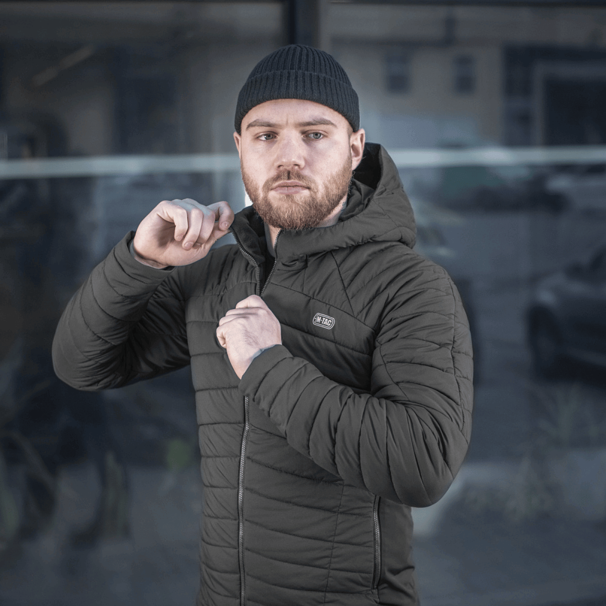 Mand iklædt M-Tac Jarl Jakke foran bygning, opkvikker jakken og ser betragtende fremad i urban omgivelse.